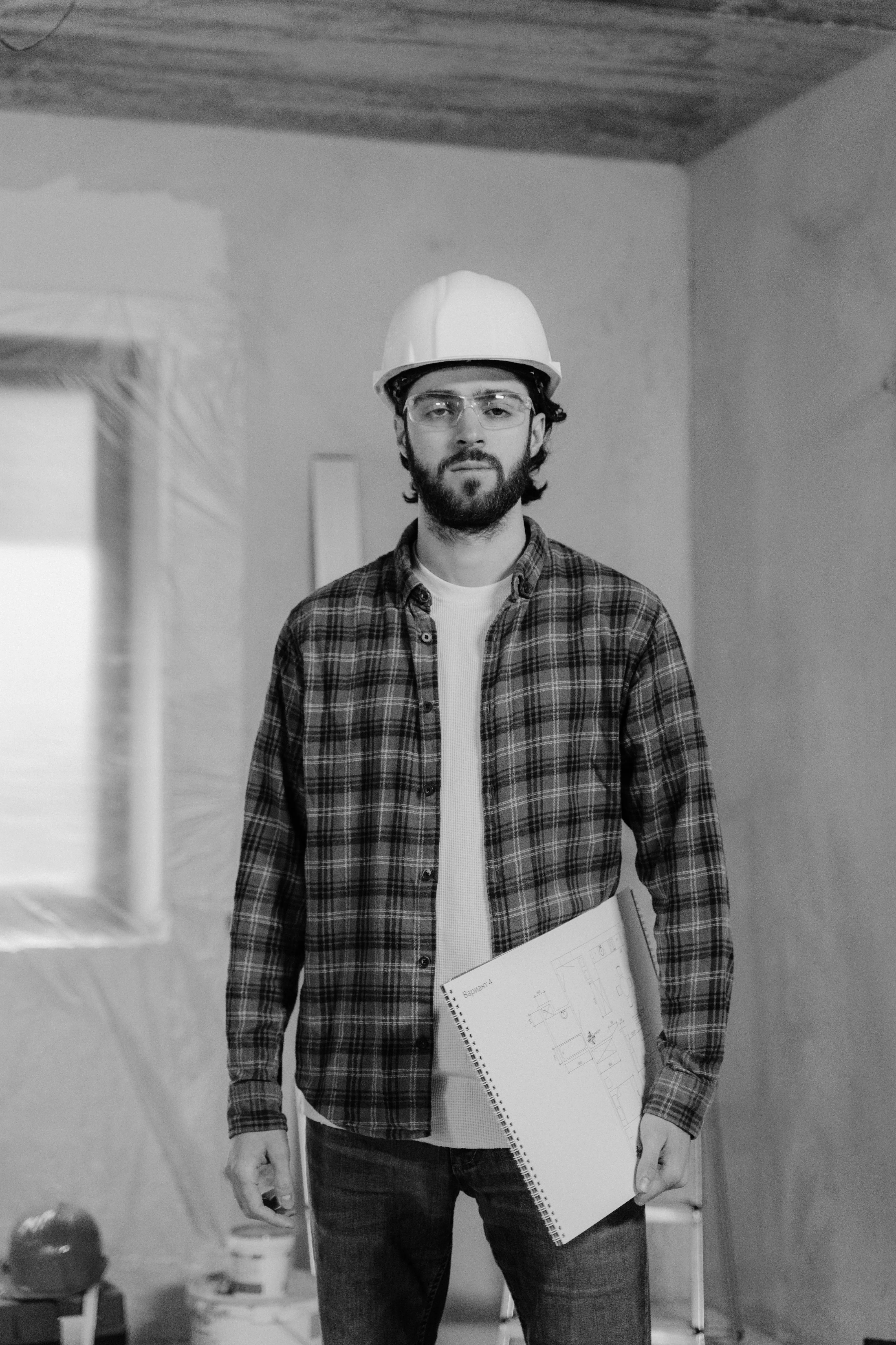 Man wearing a hard hat and safety glasses holding a blueprint in an unfinished room under construction.