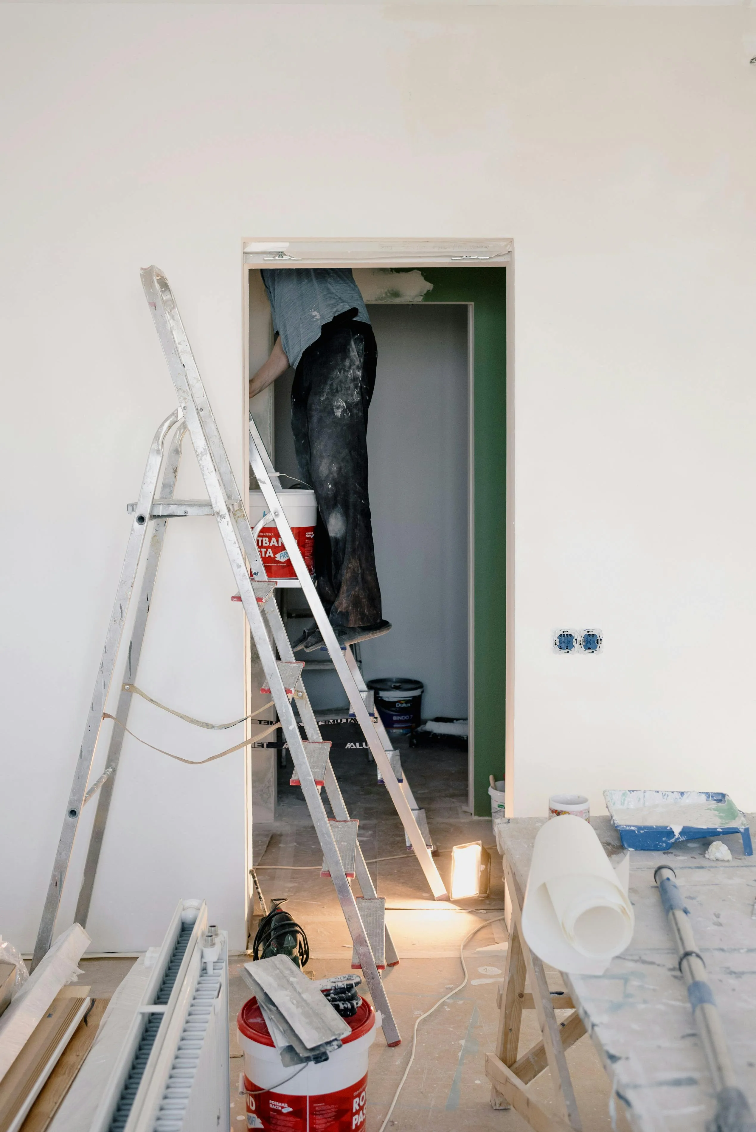 Person standing on ladder working on a doorway in a room under renovation with paint buckets and tools around.