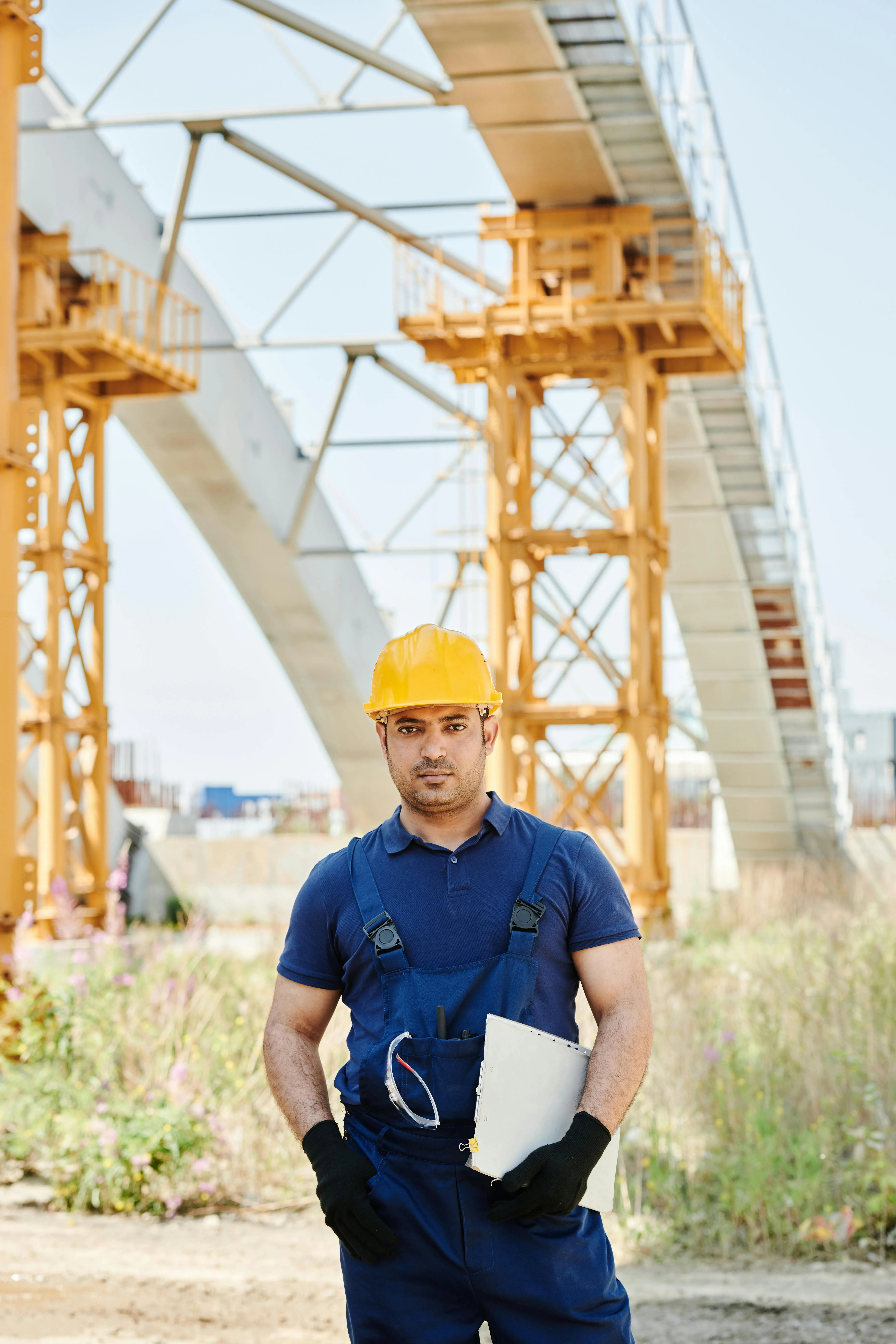 Construction worker wearing a yellow hard hat and blue overalls holding a clipboard at a construction site with metal framework in the background.