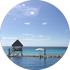 Infinity pool overlooking a calm ocean with a wooden pier, thatched hut, and white umbrella under a partly cloudy sky.