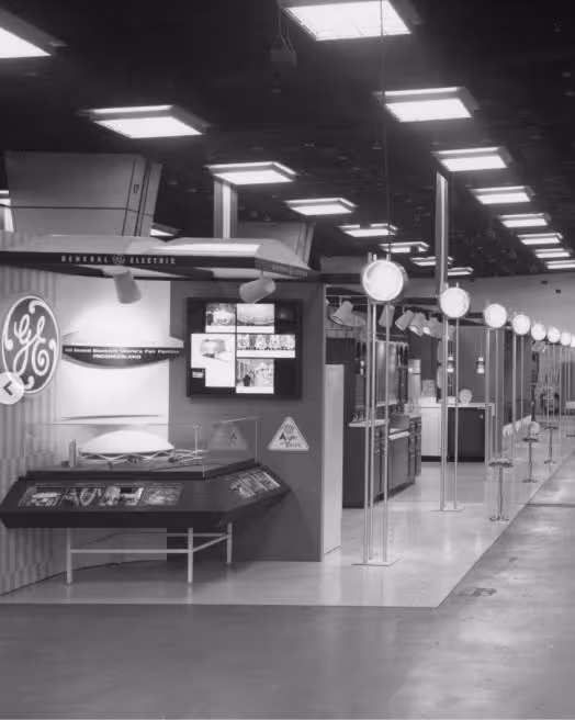 Black and white image of a General Electric exhibit with displays, signs, and overhead lights in a large room.