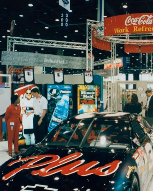 Black race car with red 'Plus' logo on hood at Coca-Cola exhibit with people in background.