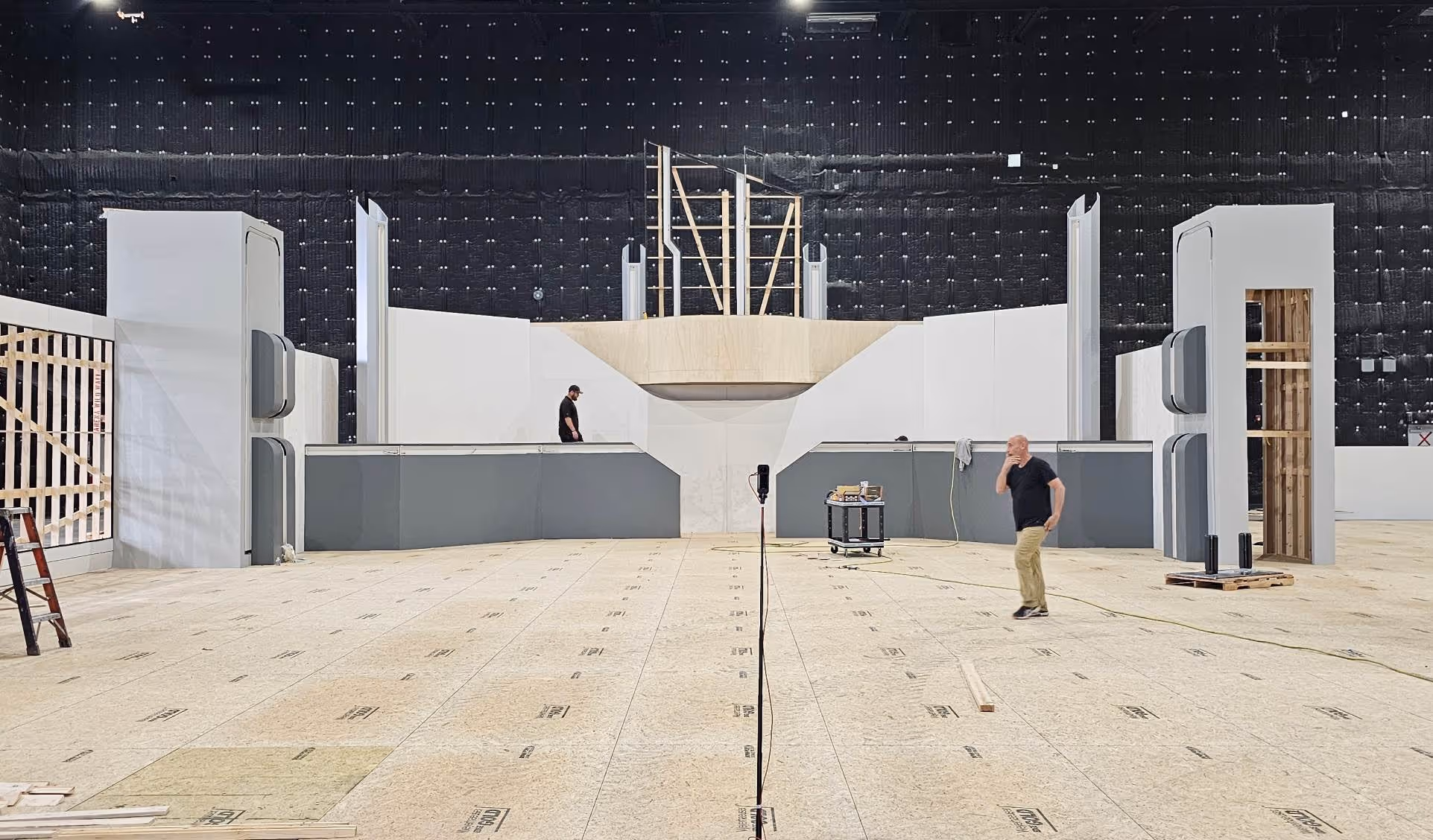 Indoor stage under construction with two men, wooden panels, and black soundproof wall background.