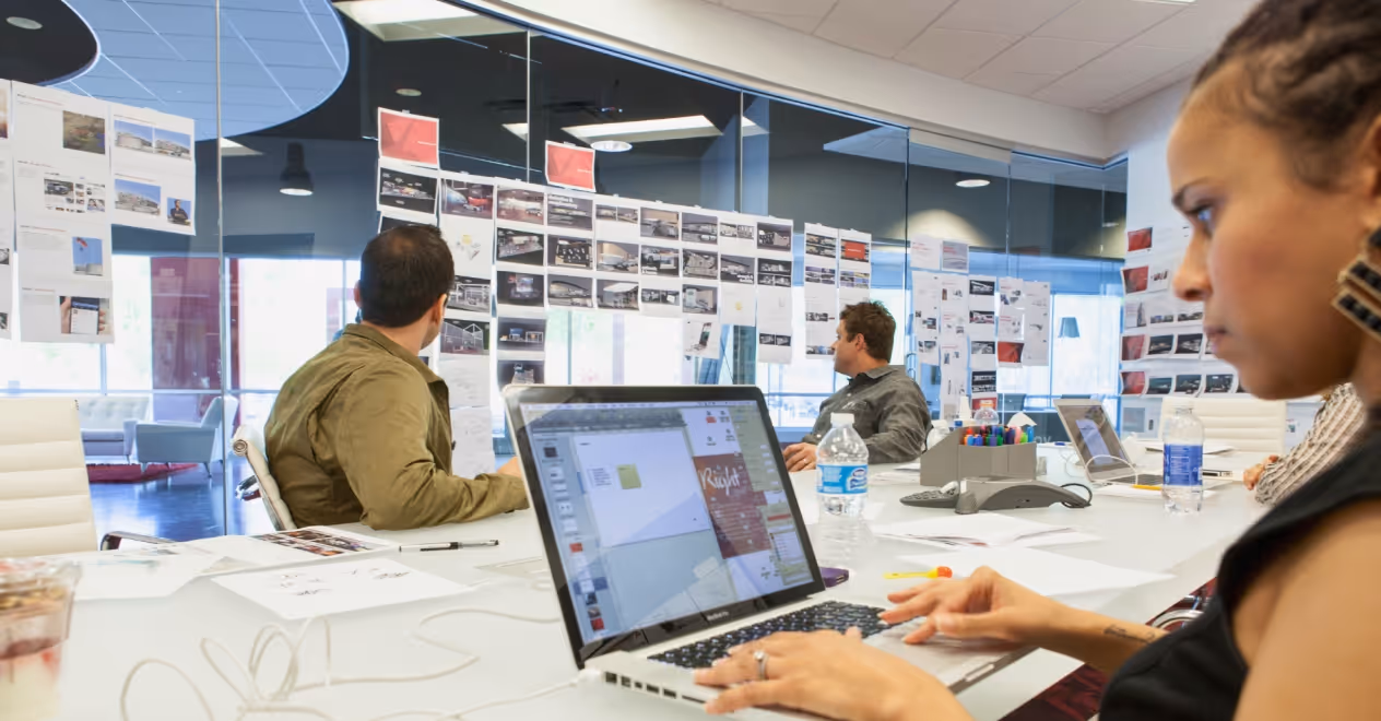 A team in a modern office reviews design layouts pinned on glass walls during a creative meeting.