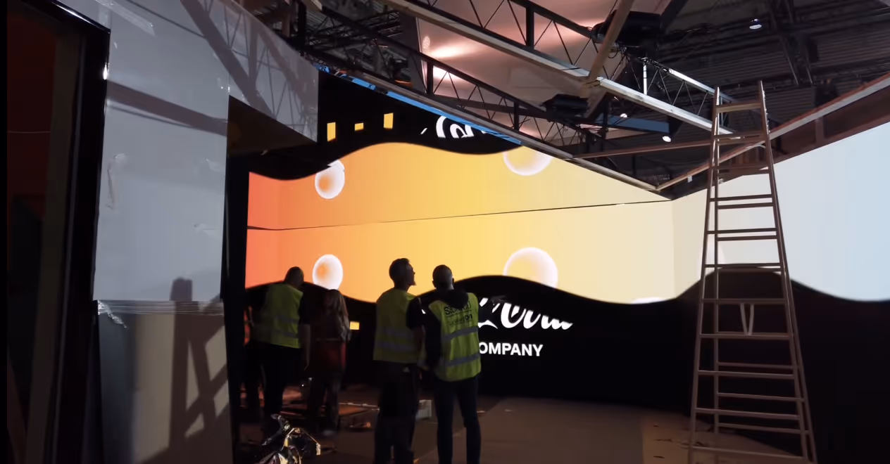 People in safety vests install a large curved digital screen display in an indoor venue with scaffolding and a ladder.