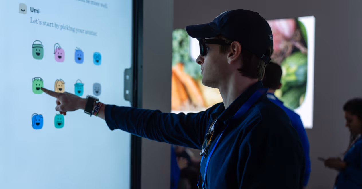 Man wearing sunglasses and a cap interacts with a touchscreen displaying colorful emoji avatars.