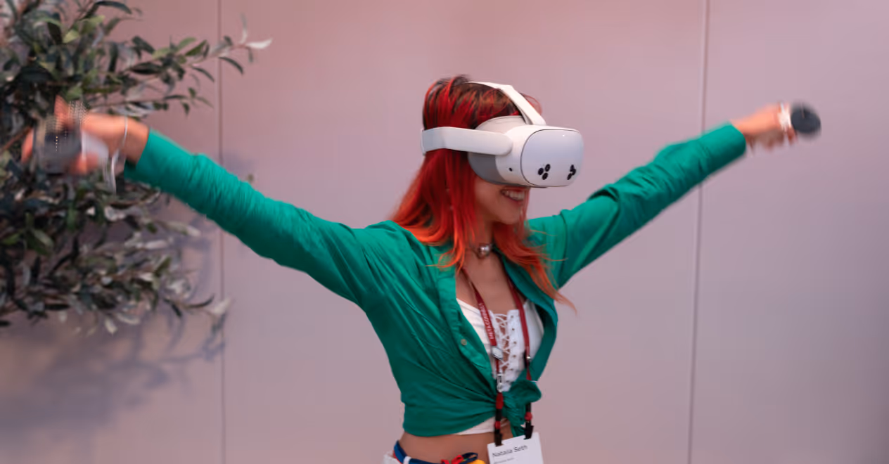 Woman with red hair wearing a green shirt using a VR headset with arms outstretched indoors.