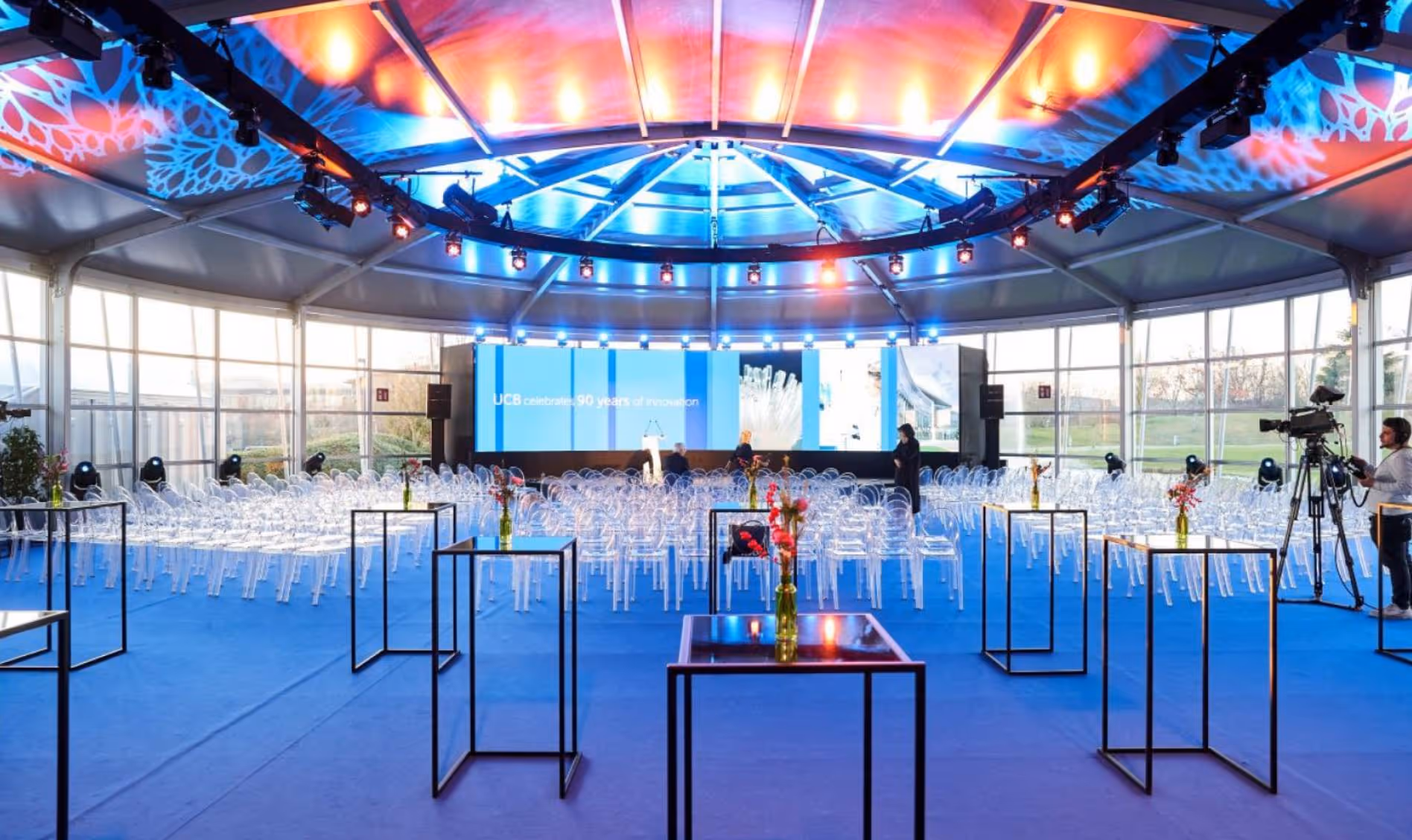 Bright event space with rows of transparent chairs, lit ceiling, stage, and camera operator.