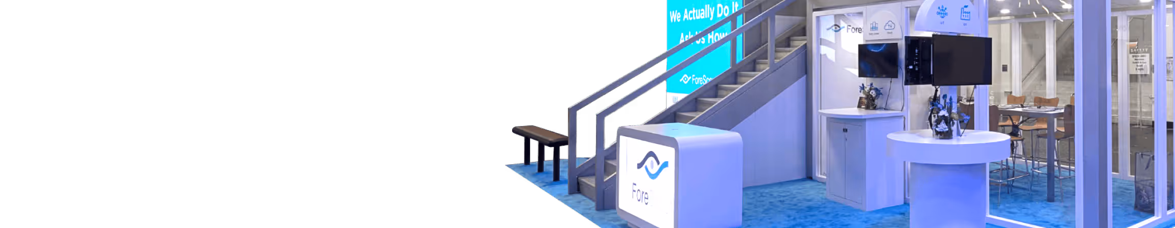 Trade show booth with ForeScout branding, monitors, a staircase, and seating area on blue carpet.