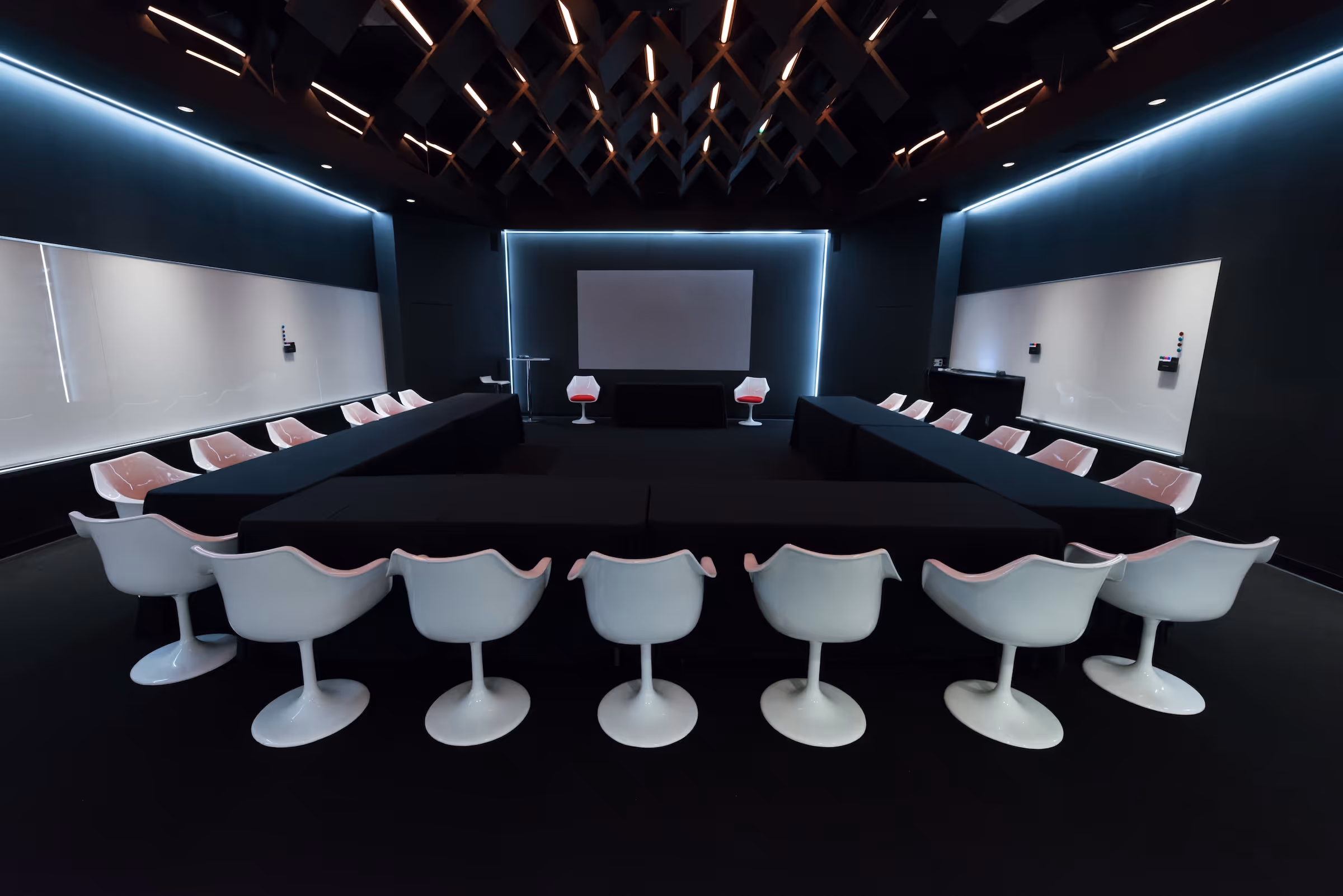 Modern dark conference room with U-shaped black tables and white swivel chairs facing a blank screen.