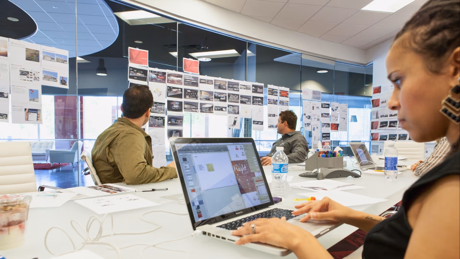 People collaborating in a meeting room with a laptop and papers, reviewing content on a glass wall.