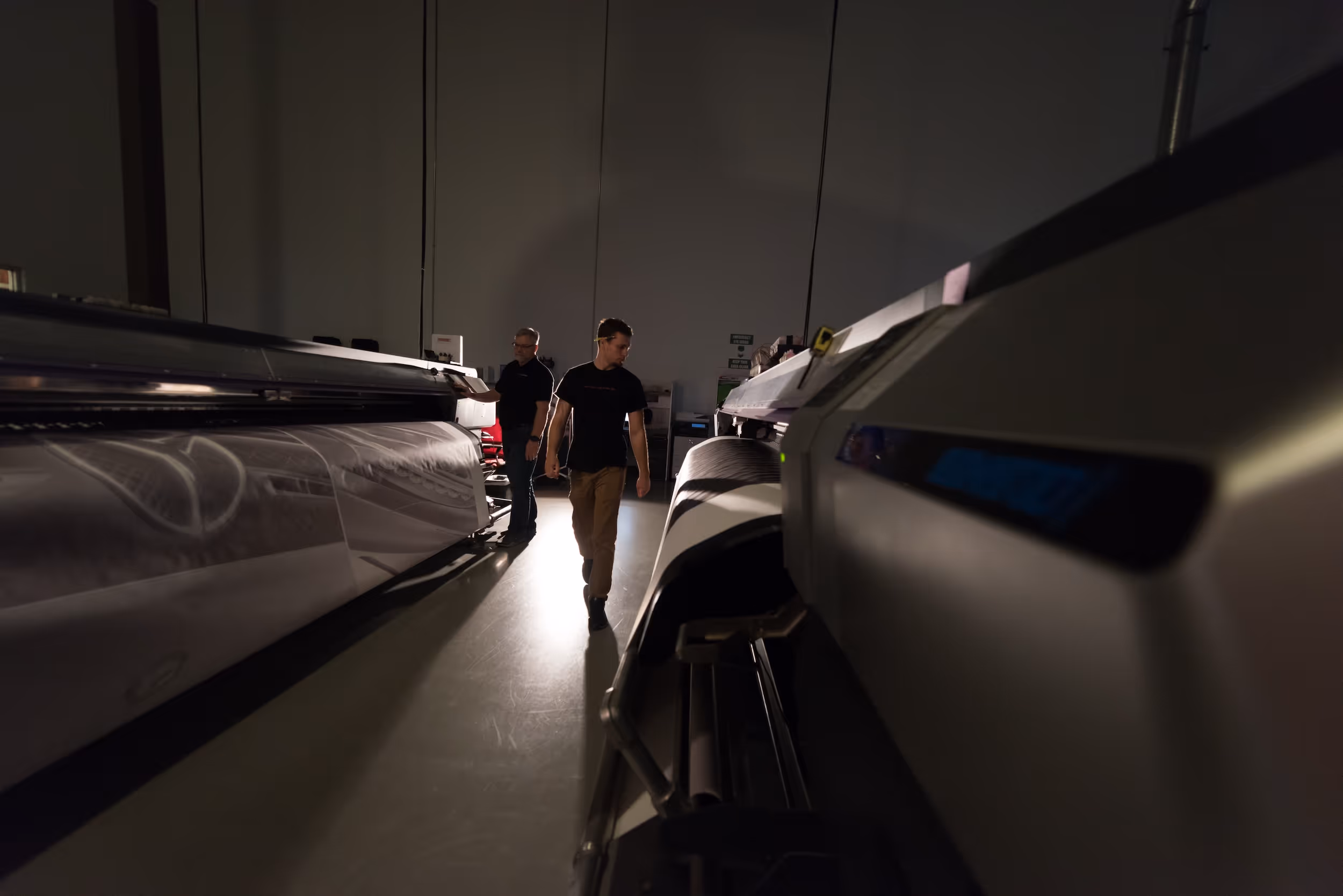 Two men operating large format printers in a dimly lit industrial printing room.