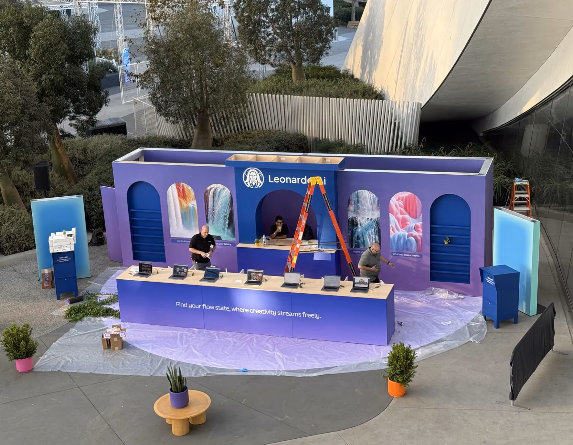 Outdoor purple booth with laptops, three men working, and art panels at a tech event setup.