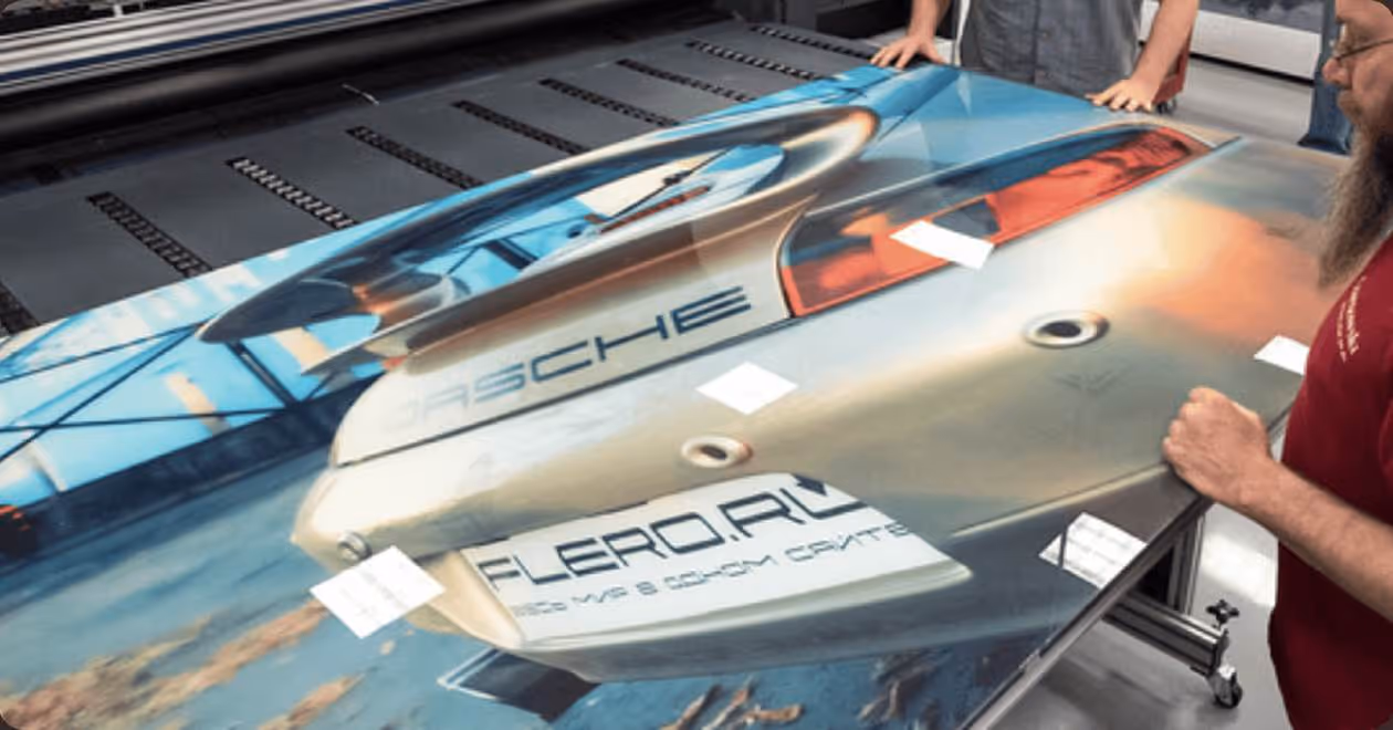 Two people inspecting a large printed image of a Porsche rear on a flat surface.