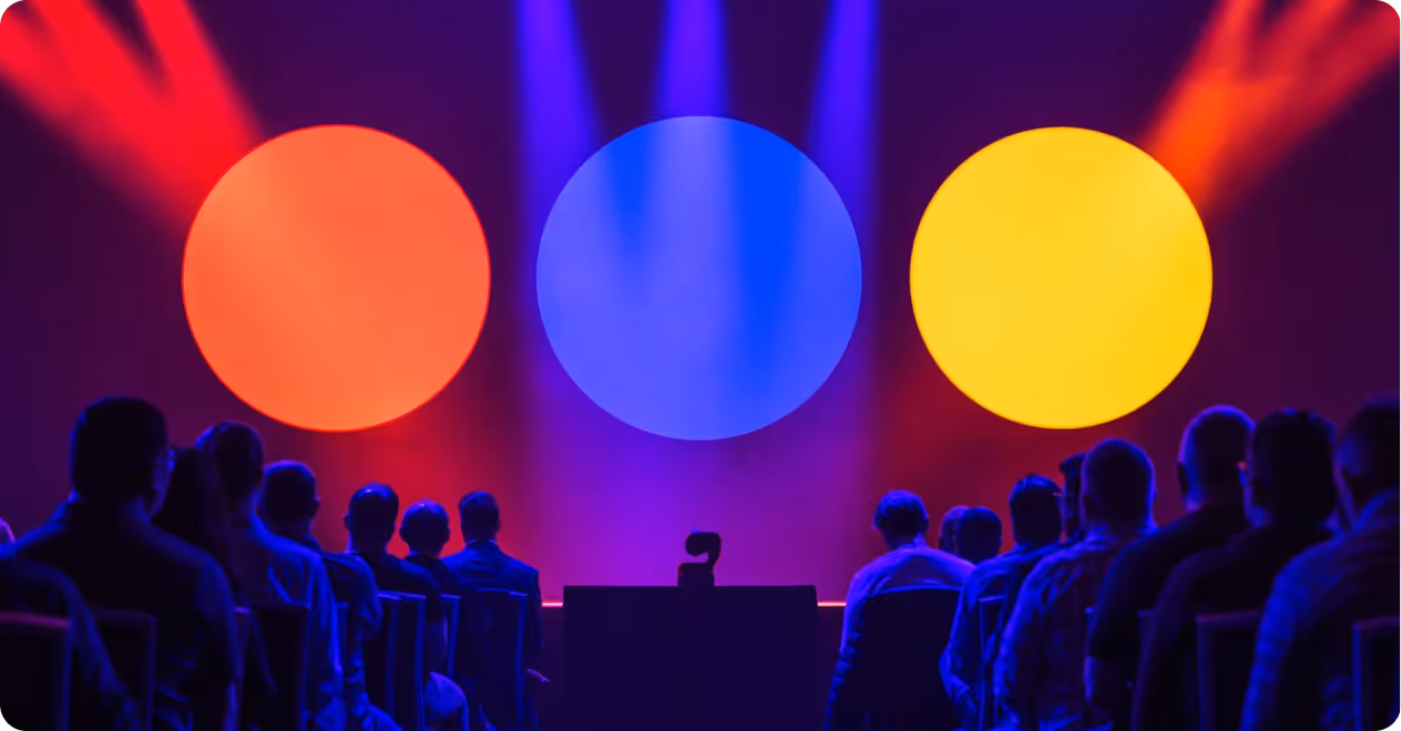 Audience seated facing a stage with large orange, blue, and yellow circular lights and colorful spotlights.