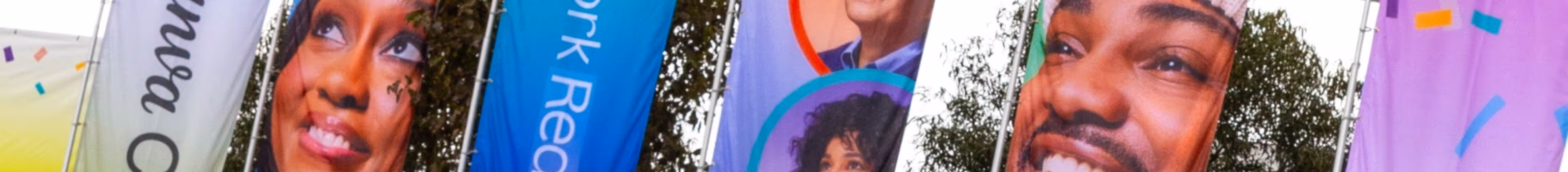 Close-up of colorful banners showing diverse smiling faces outdoors with trees in the background.