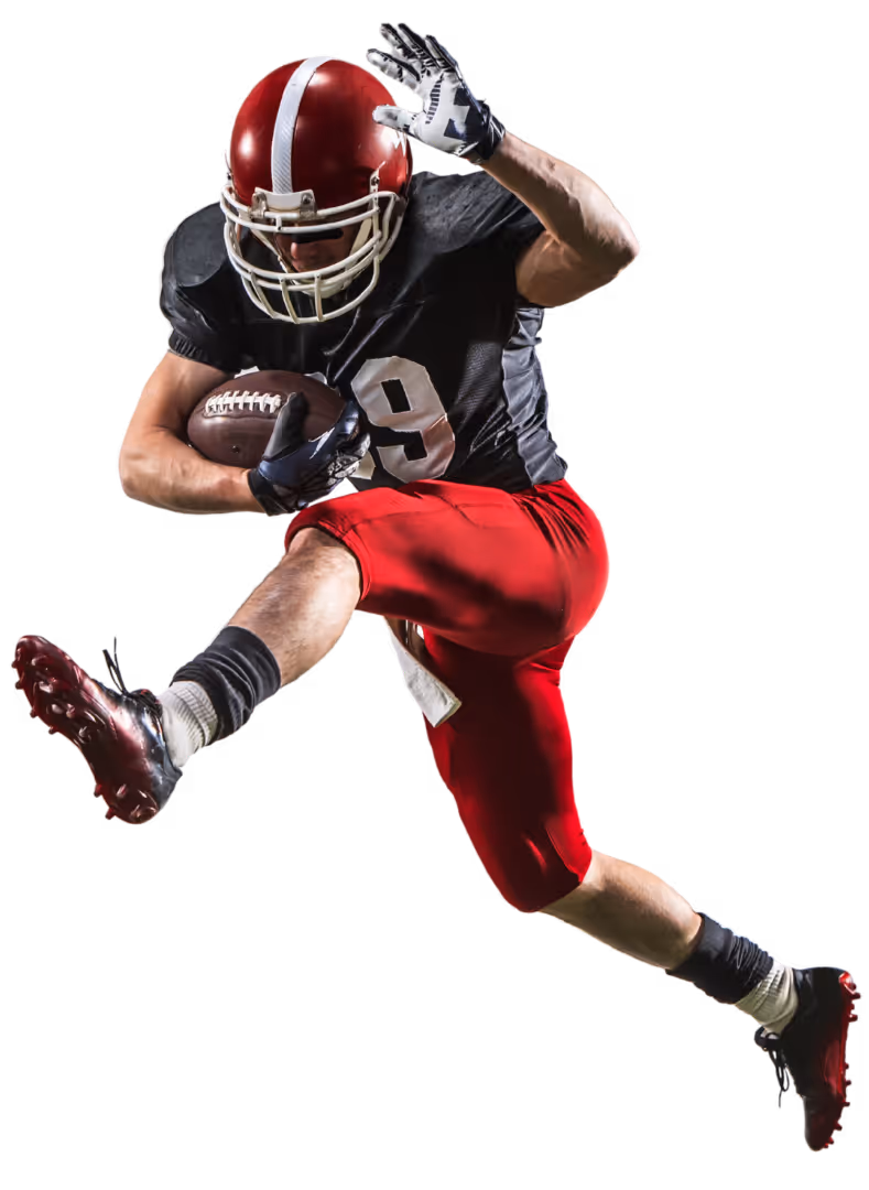 American football player in red helmet and pants, black jersey, leaping while holding a football.