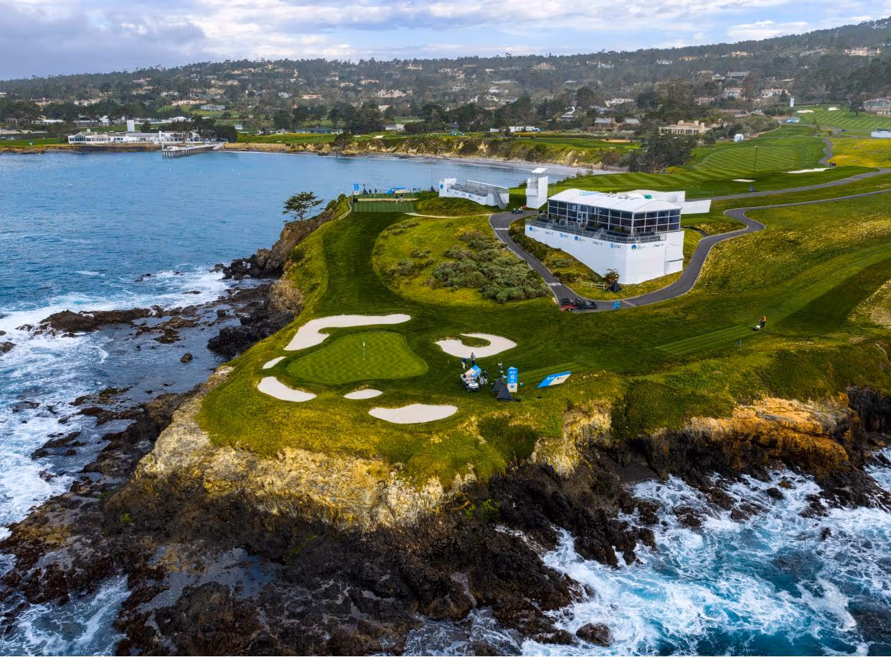 Golf course green and bunkers on rocky coast with ocean waves and event structures nearby.