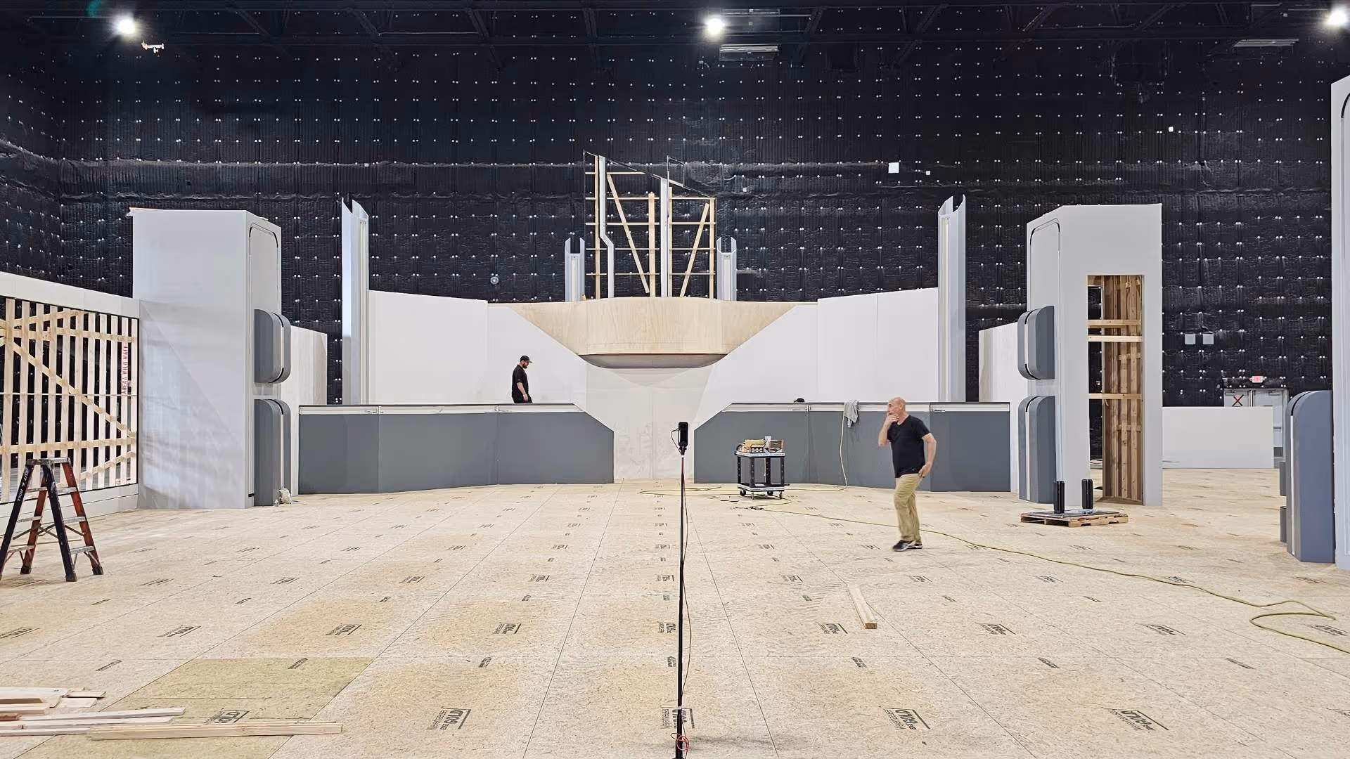 Large indoor film set under construction with plywood floor, partial walls, and two people walking.