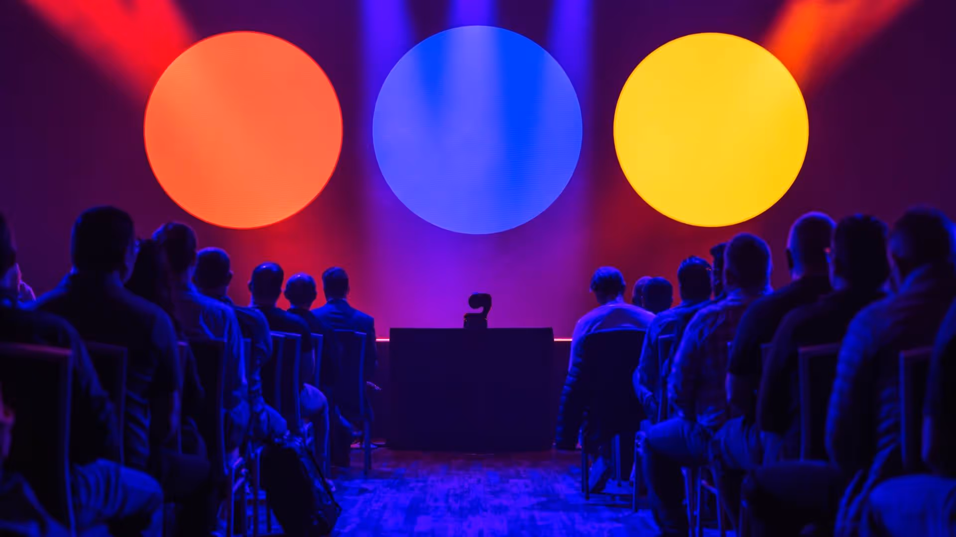Audience seated facing a stage with large orange, blue, and yellow circles and a podium with a microphone.
