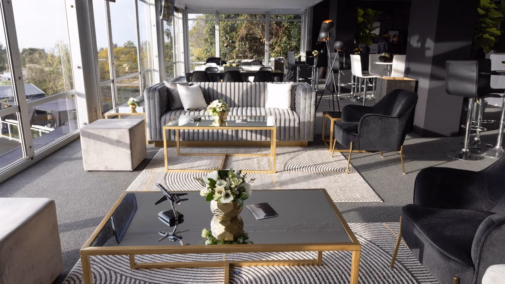 Modern lounge with gray sofa, black chairs, gold-trimmed tables, large windows, and decorative flowers.