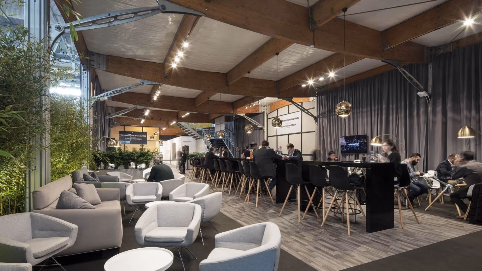 Modern coworking lounge with gray chairs, sofas, high table with black chairs, and hanging lights.