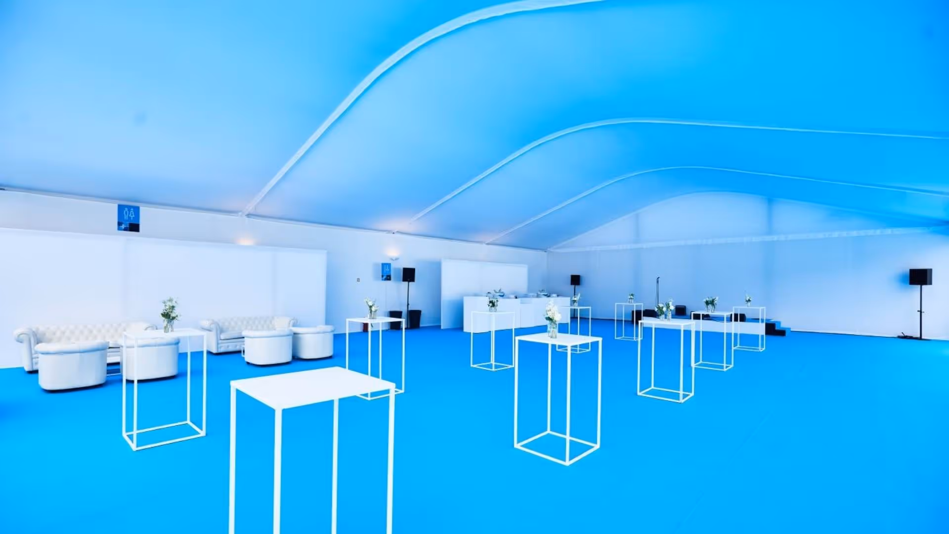Spacious event hall with bright blue floor, white high tables, sofas, and stage with speakers.
