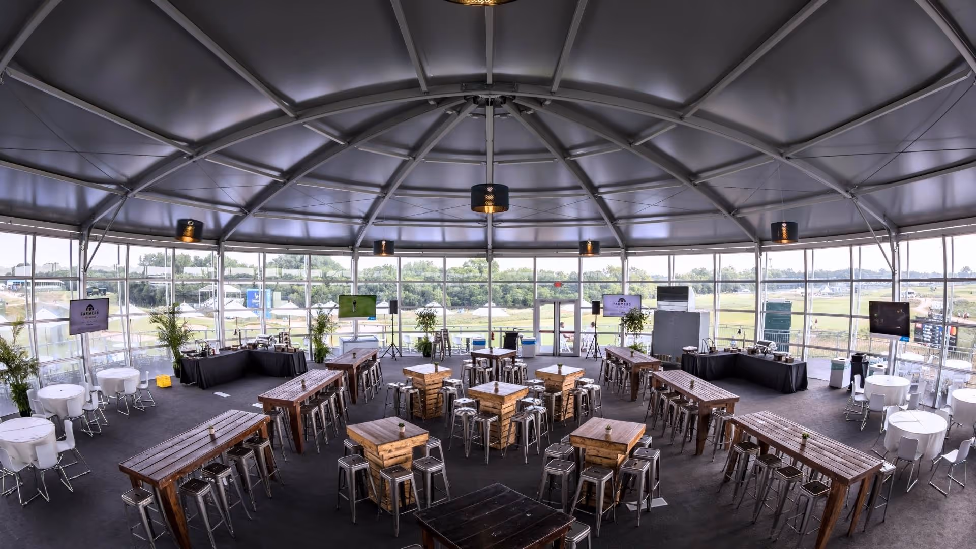 Spacious event tent with wooden tables, metal stools, round white tables, and large windows overlooking a golf course.