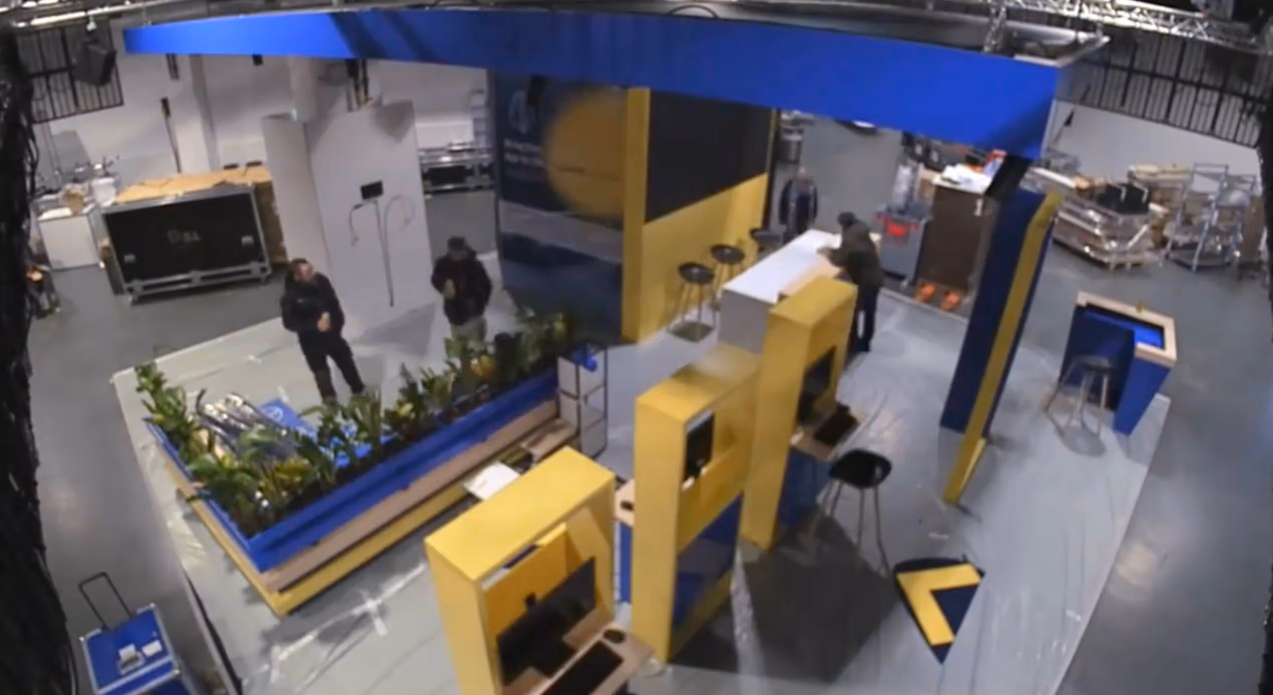 Indoor event setup with yellow and blue booths, stools, plants, and people working in an industrial space.