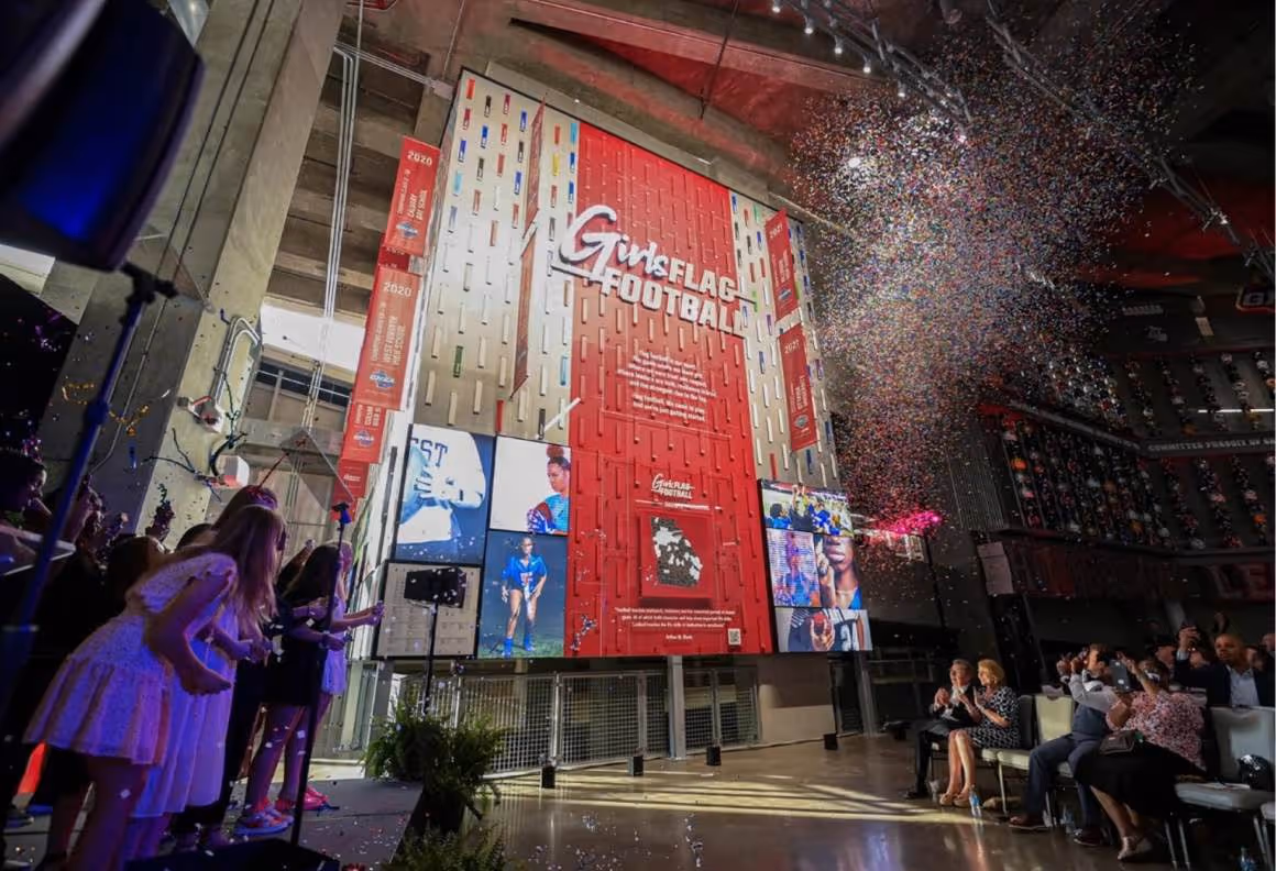 People celebrate with confetti near a large Girls Flag Football wall display with photos.