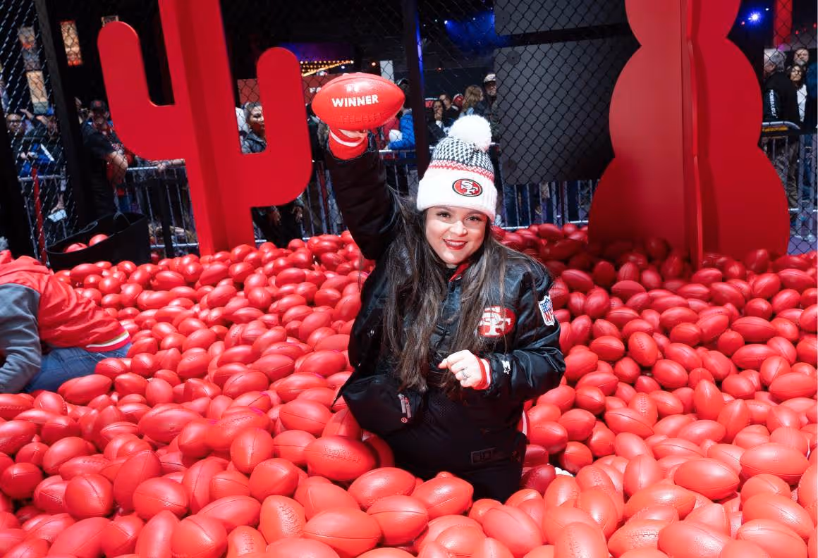 Smiling woman in San Francisco 49ers gear holding a red football in a pit filled with red footballs.