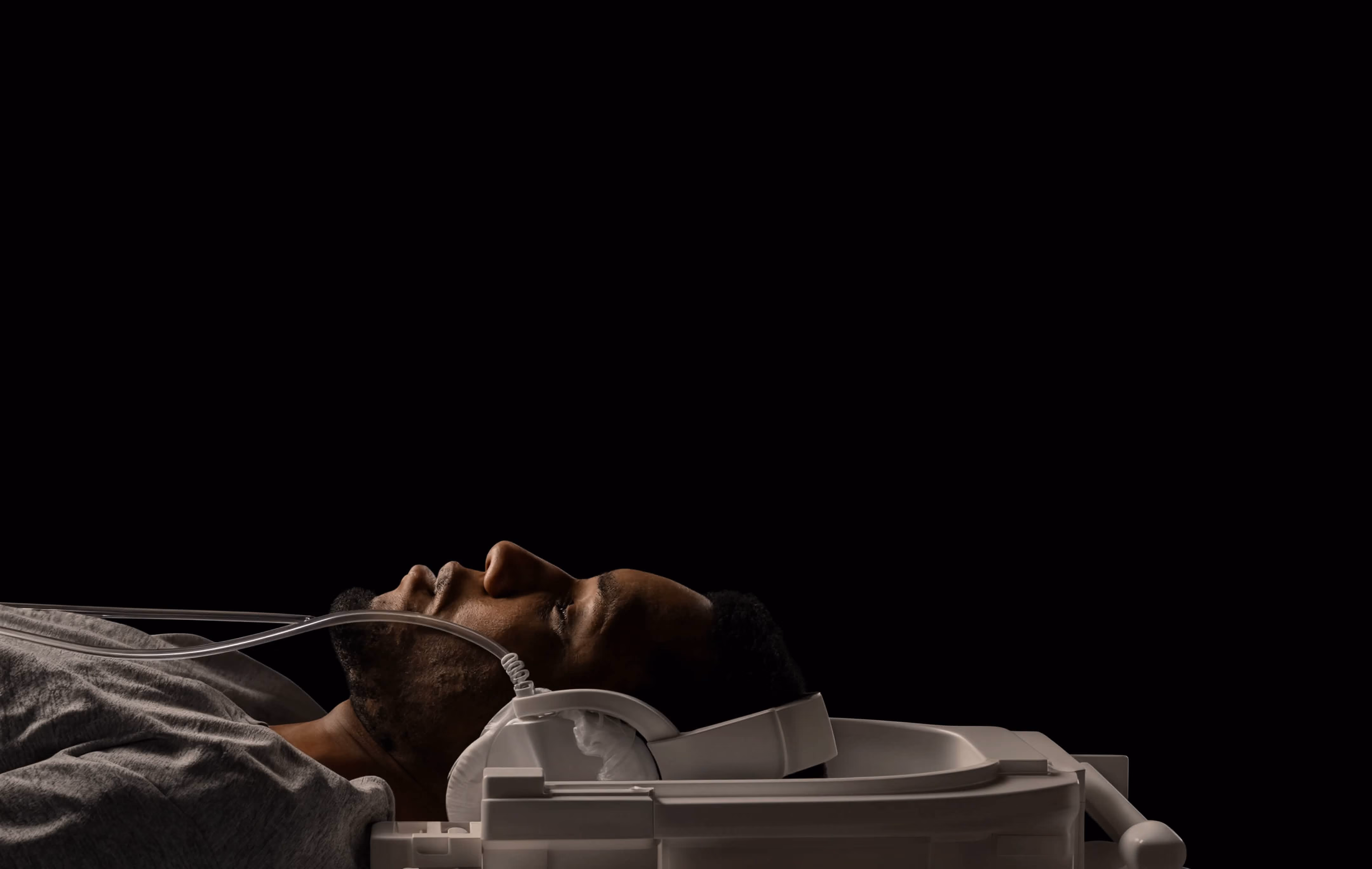 Man lying down with white headphones and nasal oxygen tubes against black background.