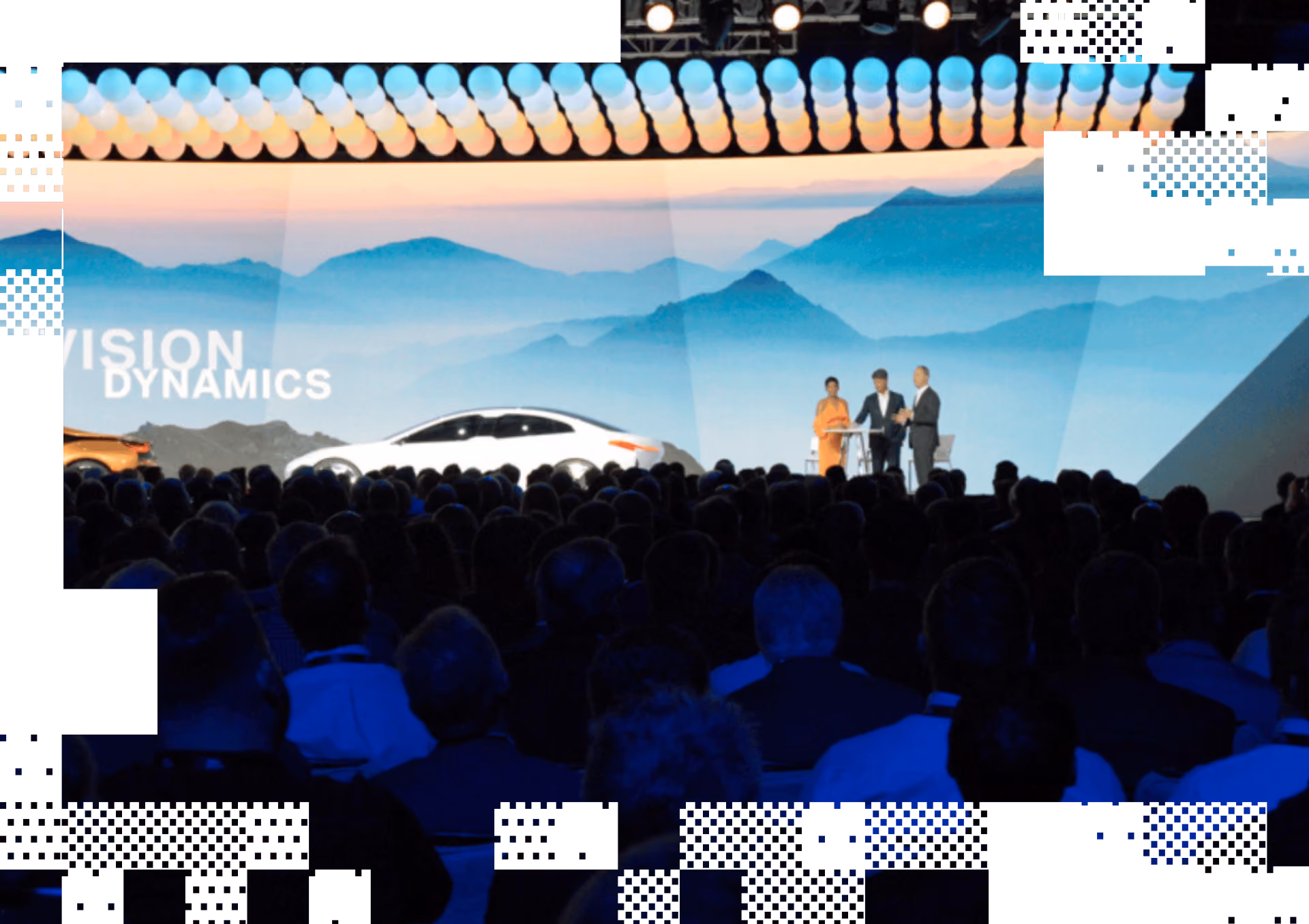 Crowd watching a presentation with two cars and three speakers on stage against a mountain backdrop.