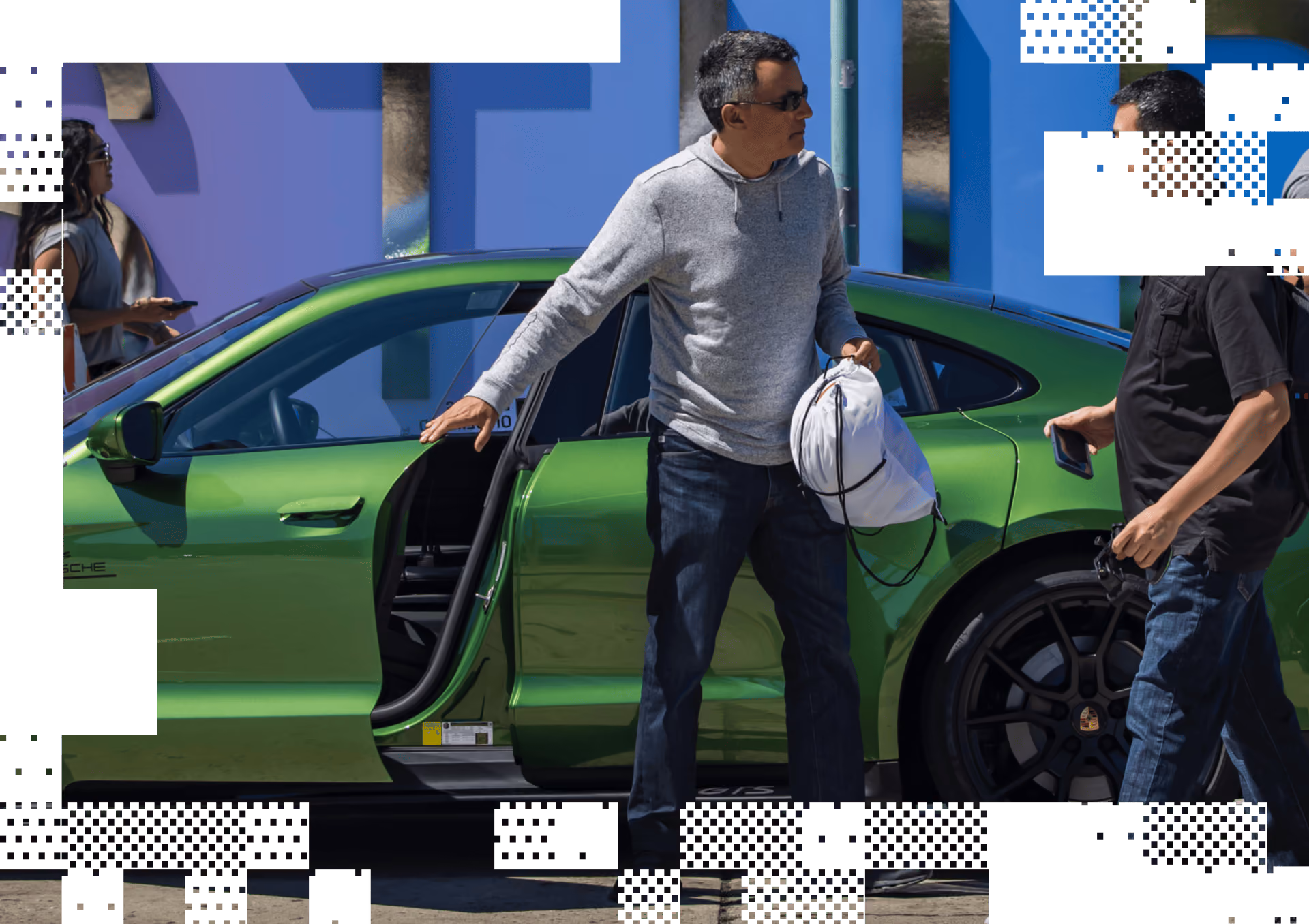 Man in gray hoodie holding a bag stands beside an open green Porsche car door talking to another man.