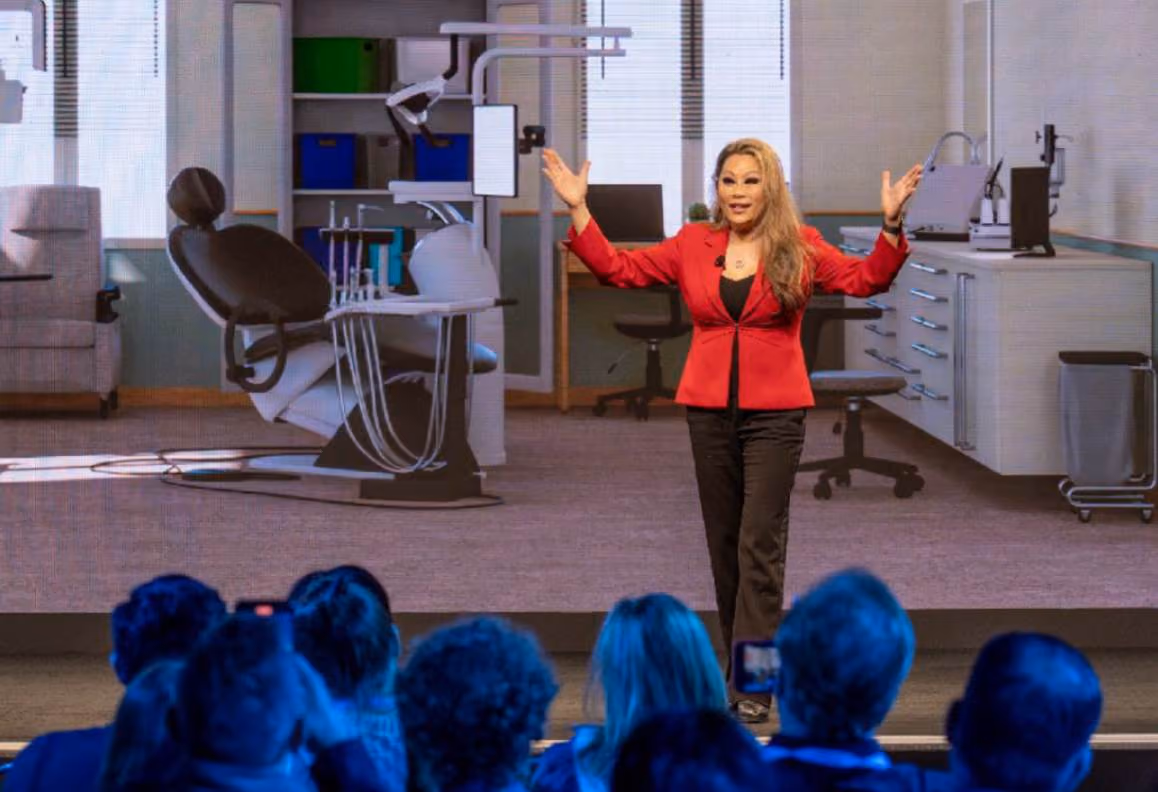 Woman in red jacket speaking on stage with dental office image projected behind an audience.