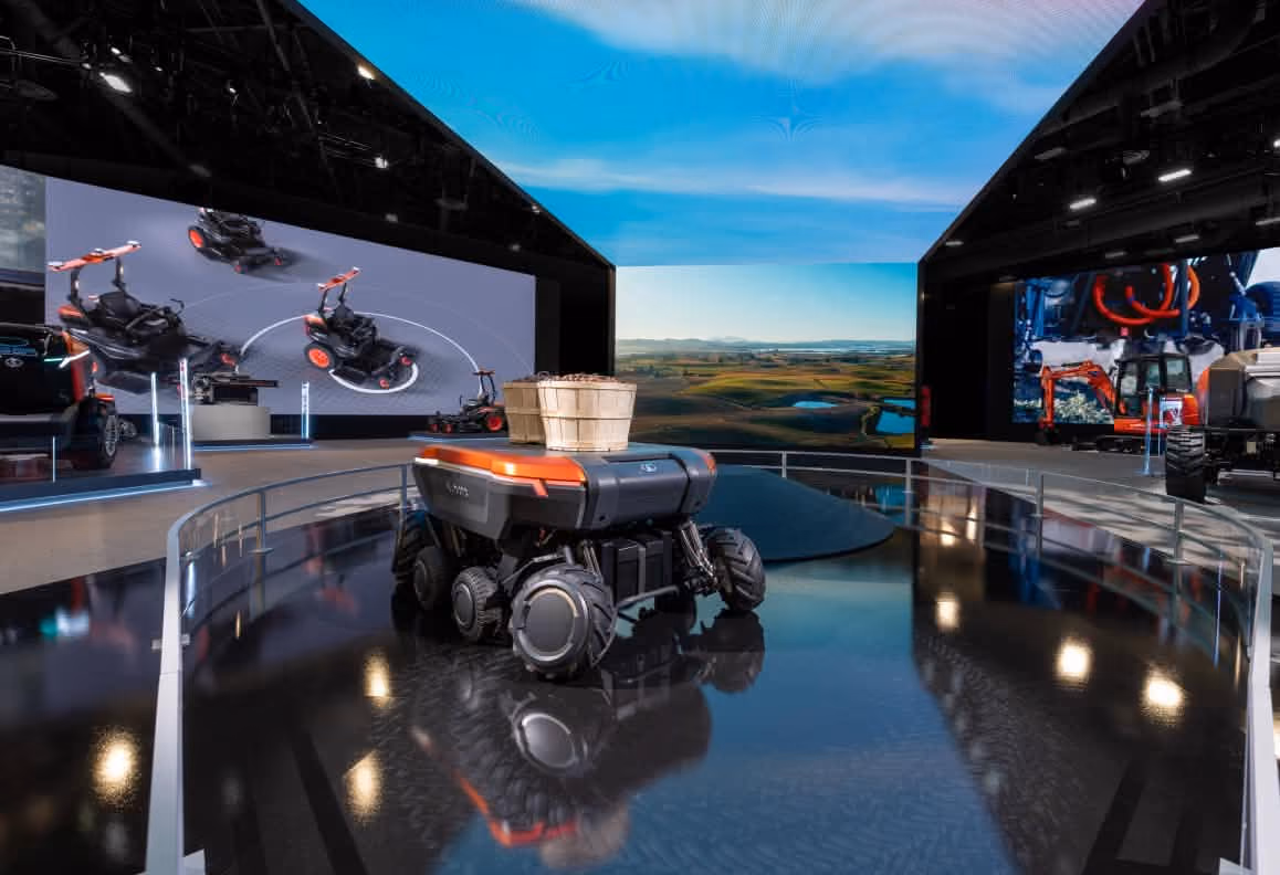 Small robotic vehicle with basket on top displayed in a modern exhibition space with large digital screens.