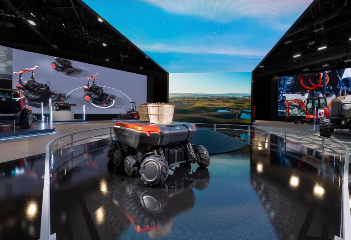 Small robotic vehicle with basket on top displayed in a modern exhibition space with large digital screens.