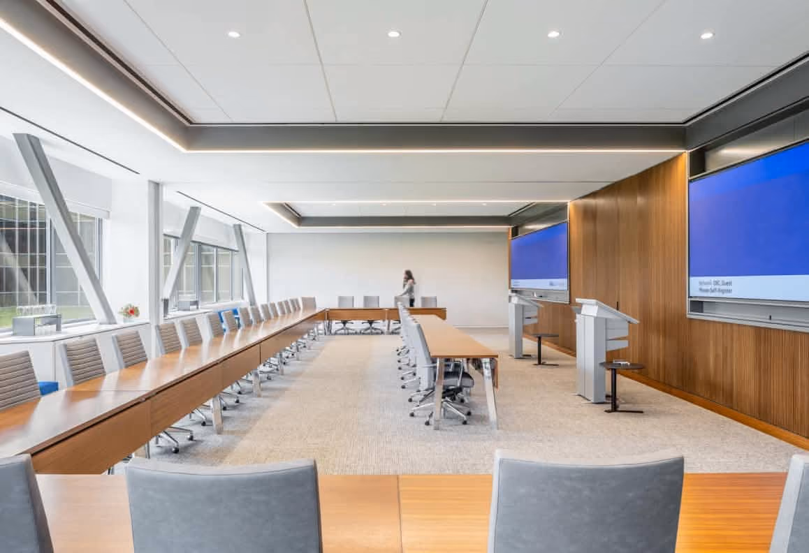 Modern conference room with long tables, swivel chairs, two large screens, and a person walking inside.
