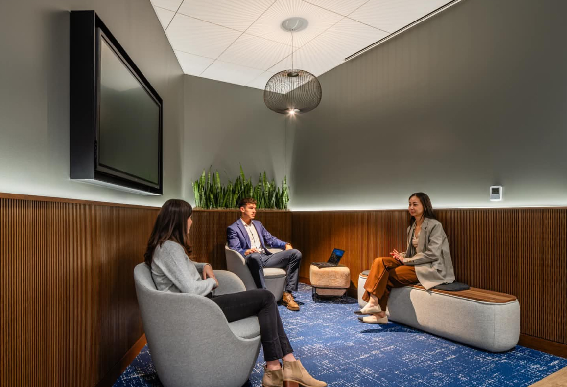 Three people having a meeting seated on modern chairs in a small office with wood paneling and blue carpet.