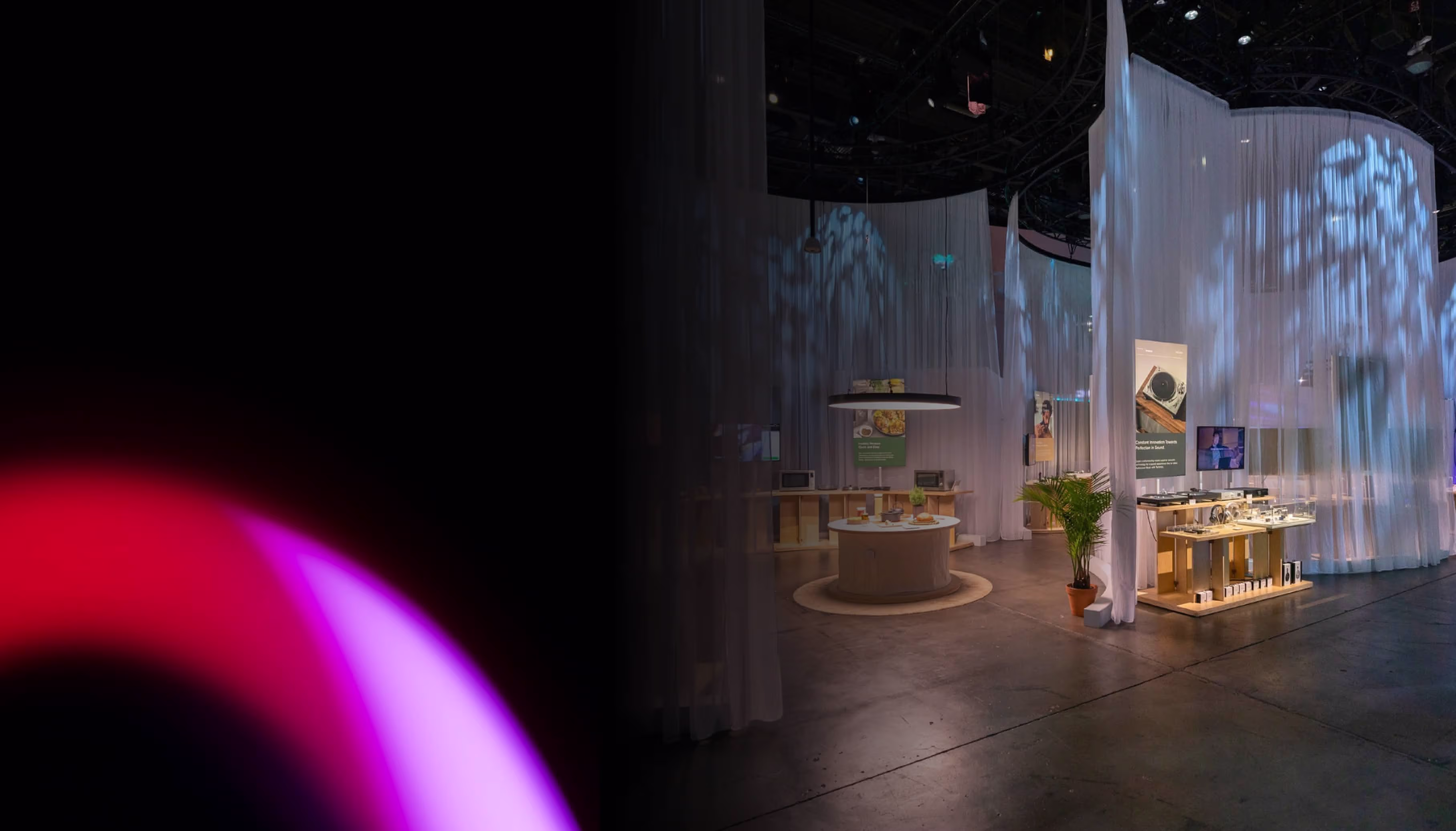 Dimly lit exhibition space with curved white translucent curtains creating partitioned areas showcasing products on round and rectangular display tables, with soft blue lighting patterns on the curtains.