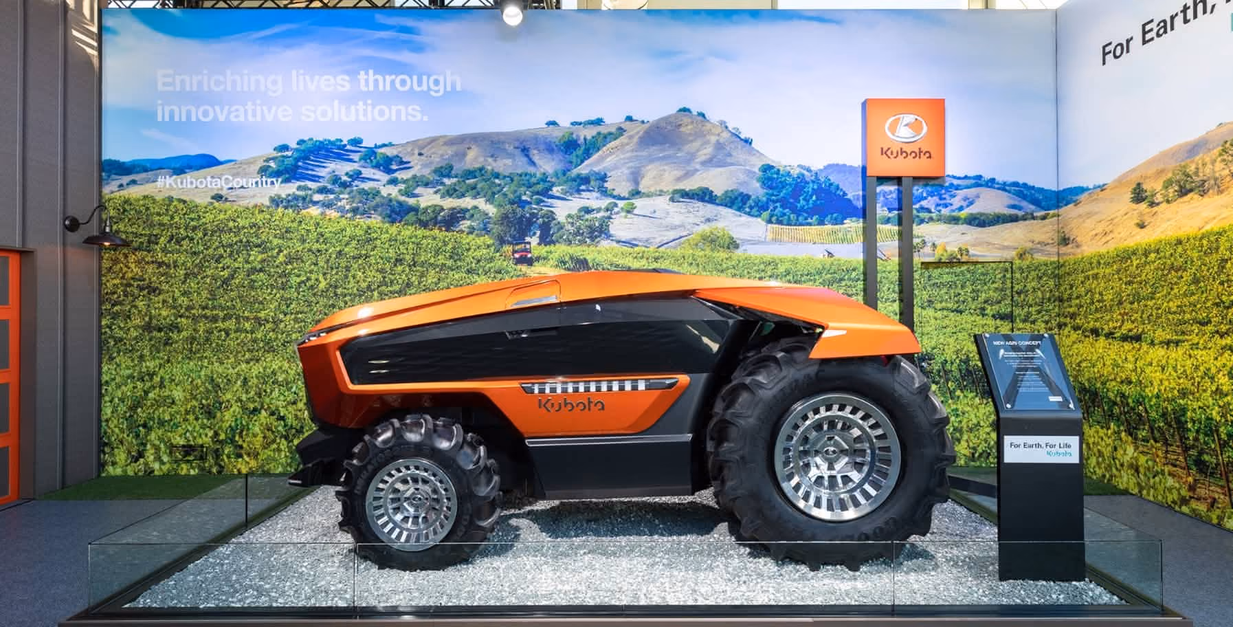 Orange and black Kubota autonomous tractor on display indoors with large backdrop of green fields and hills, accompanied by an informational stand and a Kubota sign.