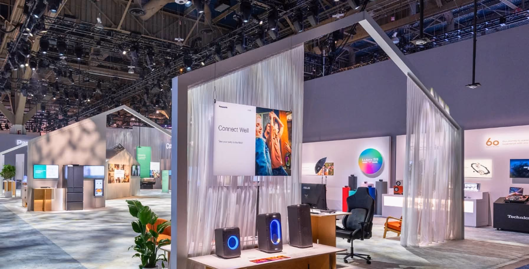 Modern exhibition booth with a hanging screen displaying 'Connect Well' and two people dancing, three speakers with blue lights on a table, a gaming chair, desk setup, and sheer white curtains creating semi-private spaces.