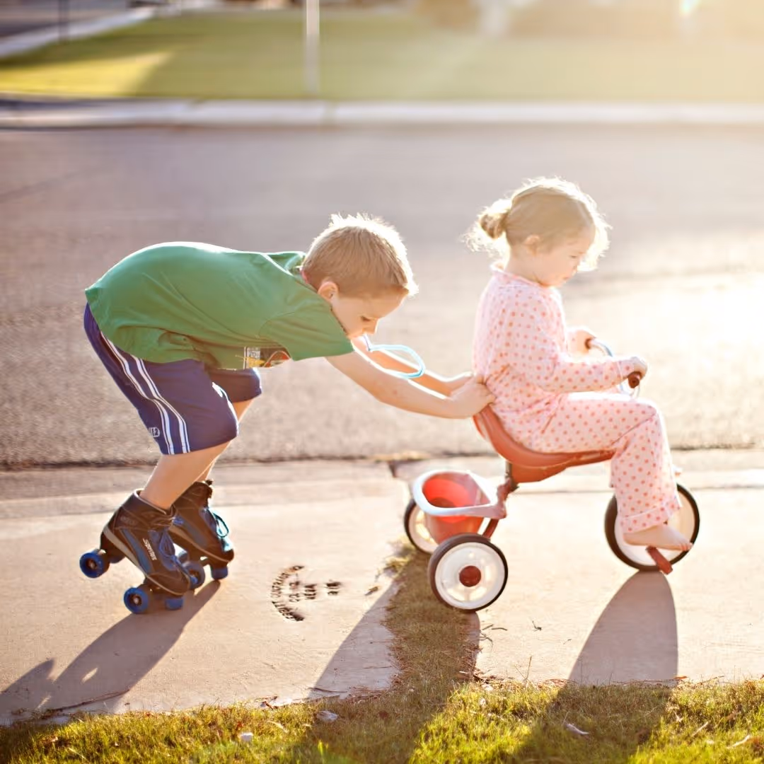a boy in rollerskates holding on the the back of a trike a little girl is riding