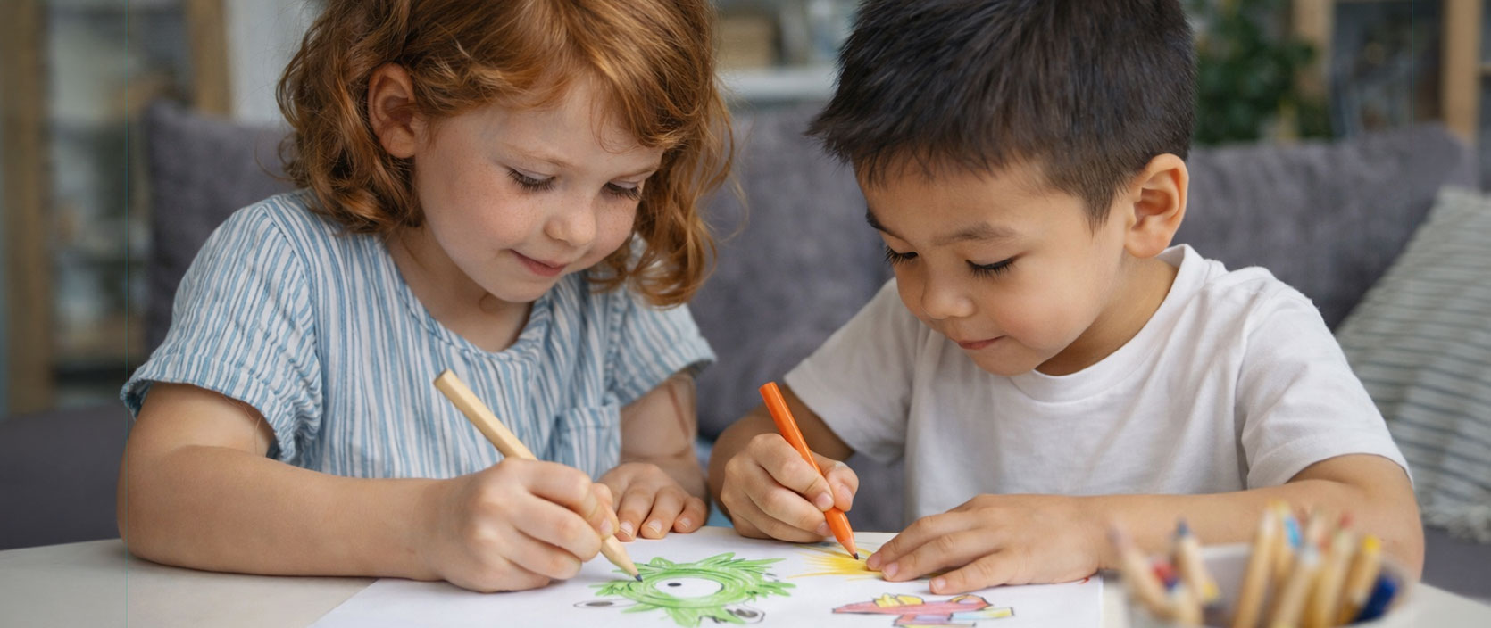Two people coloring a green cartoon character drawing on a white sheet with colored pencils on a white table.