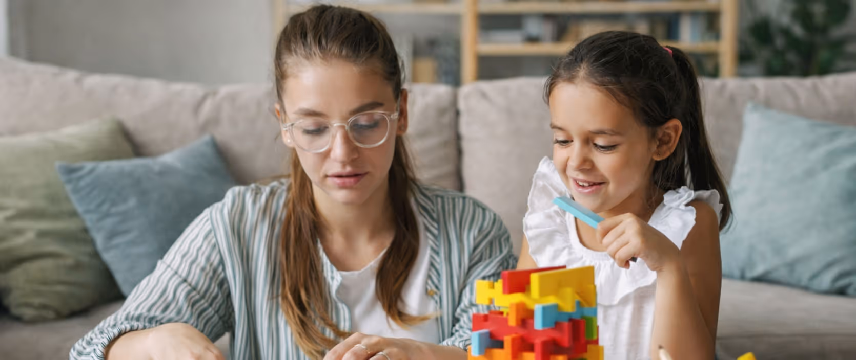 Woman and young girl sitting at a table playing with colorful building blocks and craft supplies in a living room.