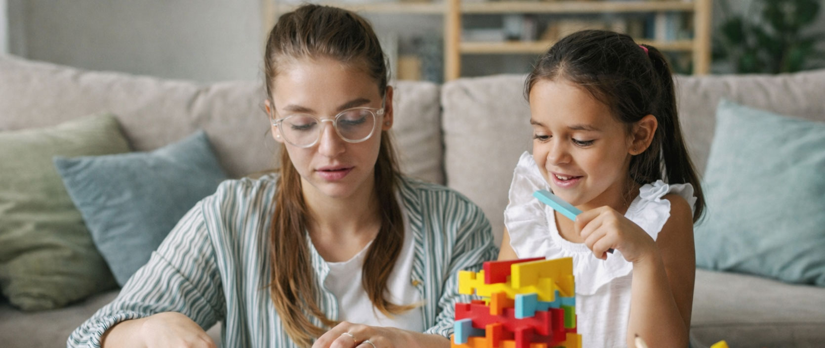 Woman and young girl sitting at a table playing with colorful building blocks and craft supplies in a living room.