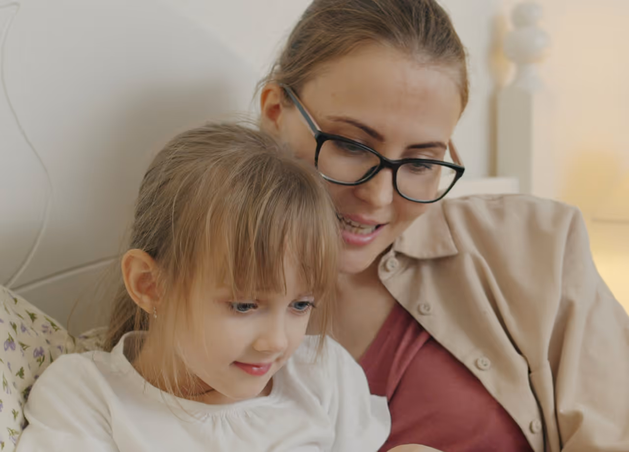 Woman wearing glasses and a beige shirt sitting closely with a young girl with bangs, both looking at something off-camera.