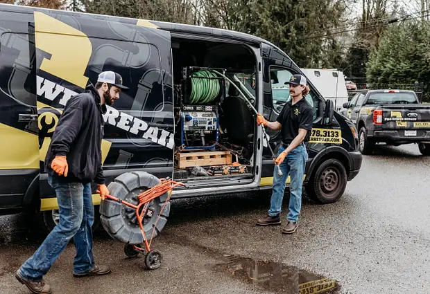 Pest control technician applying targeted tick spray treatment along shaded garden bed in residential backyard.