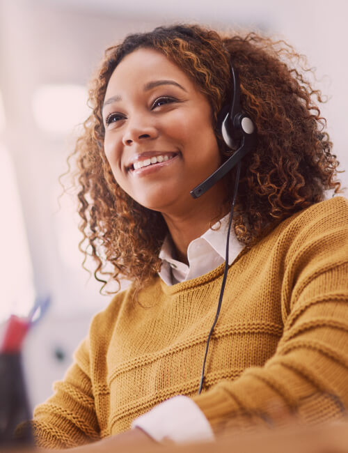Smiling woman with curly hair wearing a headset and mustard sweater in a bright office.