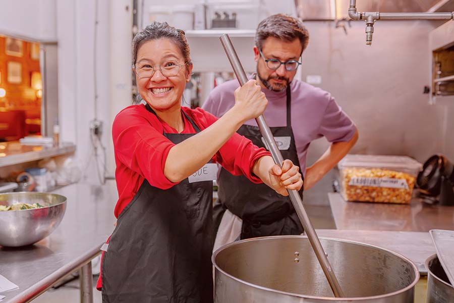 Two people in aprons cooking in an industrial kitchen, one smiling woman stirring a large pot while a man watches.